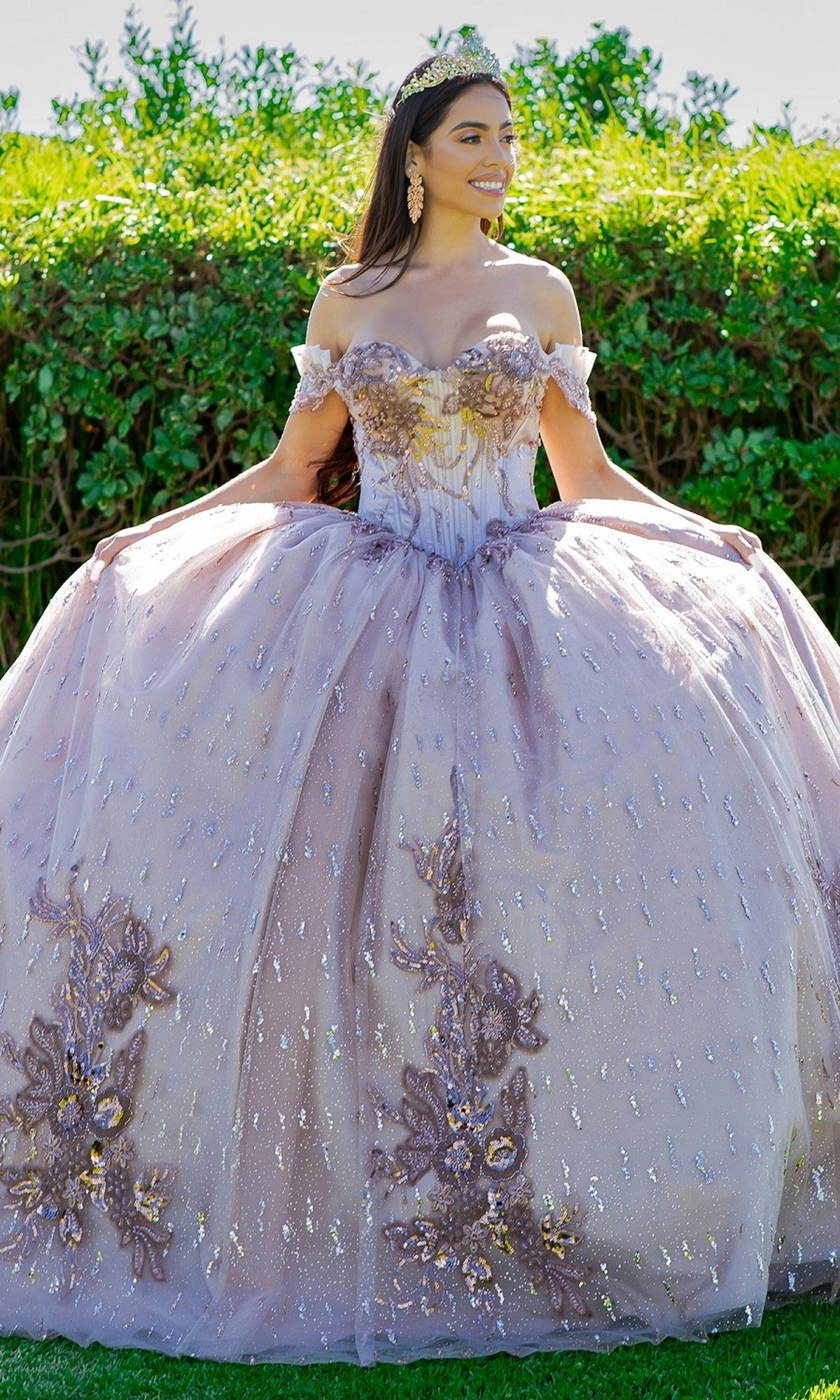 A young woman poses outdoors in the Quinceanera Ball Gown 8087J, a glitter tulle dress with off-the-shoulder sleeves and a tiara, gracefully holding out the sides of her ornate gown.